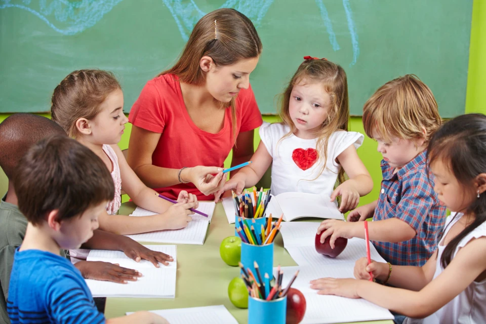 Eine Erzierherin sitzt mit mehreren Vorschulkindern gemeinsam in einer Runde an einem Tisch. Die Kinder haben Schreibhefte, Buntstifte und Äpfel. Die Erzieherung erklärt einem Kind etwas. 